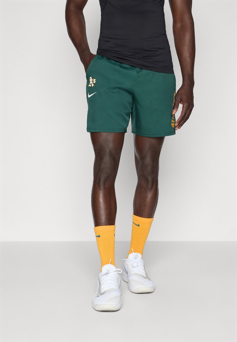 Vihreät urheilushortsit, joissa on 'A' -logo, keltaiset yksityiskohdat ja Nike-logon vaino, yhdistettynä oransseihin sukkiin ja valkoisiin lenkkikenkiin.