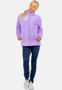 Veste en polaire violet avec fermeture éclair sur le devant, col haut et deux poches latérales. Texture douce et coupe décontractée, associée à un jean bleu et des baskets blanches.
