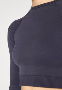 Tamno mornarsko plava majica dugih rukava izrađena od glatkog, rastezljivog materijala. Ima rebrasti rub i suptilne detalje šivanja u blizini dekoltea i rukava.