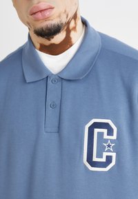 Polo en coton bleu avec un col à deux boutons, arborant un logo "C" brodé en blanc et marine sur la poitrine gauche. Tissu simple et texturé.