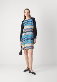 Vestido multicolorido con un patrón de zigzag en azul, amarillo y negro; de mangas largas; combinado con zapatos negros de tacón bajo y un pequeño bolso negro.