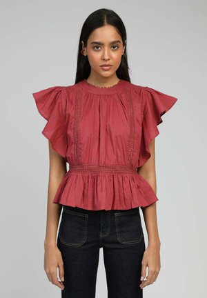 Femme aux longs cheveux foncés portant un chemisier rouge rouille à manches et taille volantées, associé à un jean foncé, se tenant devant un fond uni.