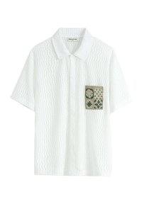 Chemise blanche à manches courtes avec un motif texturé, présentant une poche plaquée verte avec des motifs floraux et géométriques. Fermeture à boutons sur le devant.