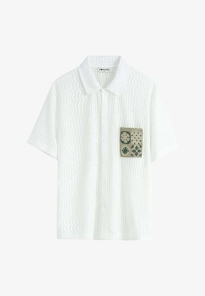 Chemise blanche à manches courtes avec un motif texturé, présentant une poche plaquée verte avec des motifs floraux et géométriques. Fermeture à boutons sur le devant.