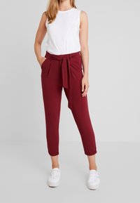 Bordeaux hoge taille broek met een ceintuur, zijzakken en taps toelopende enkellengte. Gepaarde met een witte mouwloze top en witte sneakers.