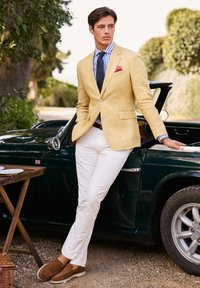 Blazer en lin jaune sur une chemise rayée bleue et blanche, pantalon blanc et mocassins marron, se tenant à côté d'une voiture classique verte.
