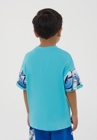 T-shirt corto turchese con stampa grafica colorata sulle maniche. Materiale in cotone con vestibilità rilassata e collo giro.