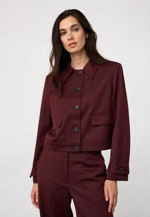 Femme aux longs cheveux foncés portant une veste bordeaux boutonnée et un pantalon assorti sur un fond clair uni.