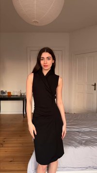 Γυναίκα με μακριά σκούρα μαλλιά που φοράει αμάνικο μαύρο φόρεμα στέκεται σε ένα υπνοδωμάτιο με ξύλινο πάτωμα, λευκό κρεβάτι, τραπέζι και φωτιστικό οροφής από χάρτινο φανάρι.