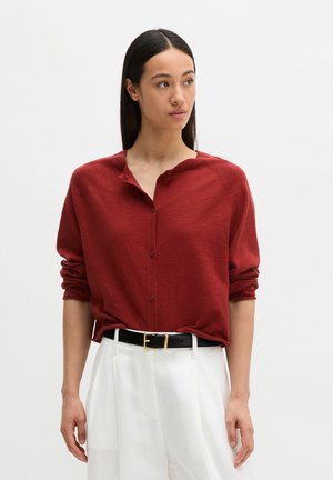 Kobieta nosząca luźny czerwony sweter na guziki z podwiniętymi rękawami, włożony do białych spodni z wysokim stanem i czarnym paskiem.