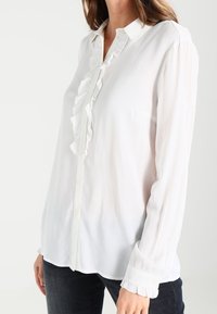 Camicia bianca a maniche lunghe con chiusura a bottoni, colletto a ruches e maniche con polsini. Realizzata in tessuto leggero e trasparente.