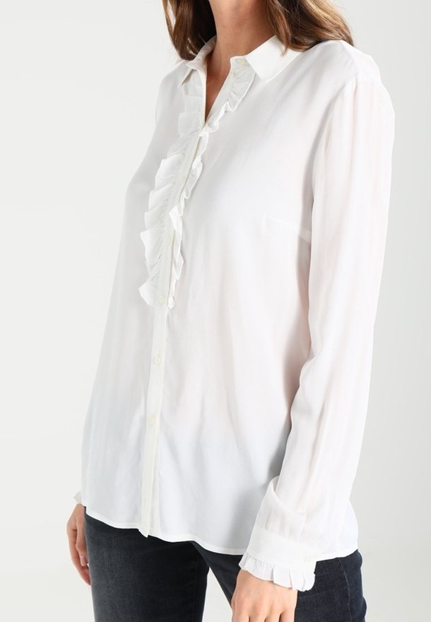 Camicia bianca a maniche lunghe con chiusura a bottoni, colletto a ruches e maniche con polsini. Realizzata in tessuto leggero e trasparente.