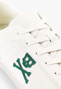 Zapatilla de ante blanca con diseño de calavera y huesos cruzados en verde. Presenta cordones planos y acentos cosidos. Superficie lisa y texturizada.