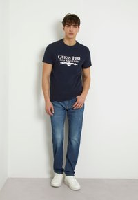 T-shirt in cotone blu navy con stampa bianca "Guess 1981 Los Angeles", abbinata a jeans blu chiaro e sneakers bianche; vestibilità casual.