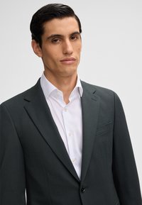 Completo su misura verde scuro con revers, chiusura a un bottone e taschino frontale, indossato sopra una camicia bianca con colletto. Tessuto dalla texture liscia.