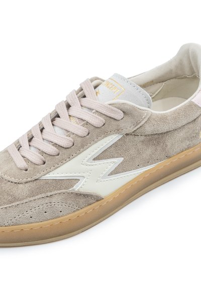 Baskets en daim beige avec un accent en zigzag blanc, des lacets beiges, une semelle gomme et un logo doré sur la languette, présentées en gros plan sous un angle.