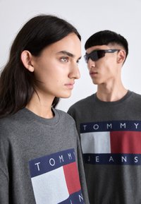 Sudadera gris con un gran logo "TOMMY JEANS" en colores bloque rojo, azul y blanco. Textura suave, cuello redondo y corte relajado.