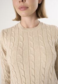 Beige gestrickter Pullover mit strukturiertem Kabelmuster, Rundhalsausschnitt und gerippten Bündchen. Der Stoff wirkt weich und leicht strukturiert.
