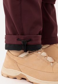 Bordeauxkleurige waterdichte broek tot de enkels met een trekkoord, gecombineerd met lichtbruine wandelschoenen met een gemêleerde leren bovenkant en rubberen zolen.