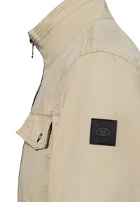 Beige Jacke mit hohem Kragen, Reißverschluss und linker Brusttasche. Verfügt über ein schwarzes Etikett mit Logo. Strukturiertes Material.