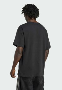Mand med sorte dreadlocks iført en simpel sort oversized t-shirt og sorte bukser med hvide striber, vist fra ryggen.