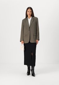 Blazer oversized vert olive, chemise blanche à col, jupe midi noire avec fente, bottes noires jusqu'aux genoux, petit sac à main crème.