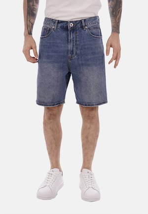 Uomo che indossa pantaloncini di jeans blu slavato di media lunghezza, sneakers bianche e una t-shirt bianca, con gli avambracci tatuati visibili.