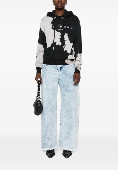 Felpa tie-dye bianca e nera con branding bianco, abbinata a jeans a gamba larga blu chiaro con orli sfrangiati e scarpe nere a punta.