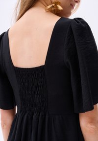 Schwarzes Kleid mit kurzen Puffärmeln, das einen quadratischen Rücken und Raffung in der Taille aufweist, aus weichem, strukturiertem Stoff gefertigt.