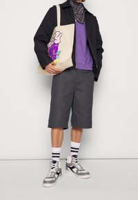 Giacca nera, camicia viola, pantaloni grigi, calze bianche con strisce nere, sneaker grigie e bianche. Tiene una borsa beige con design grafico.