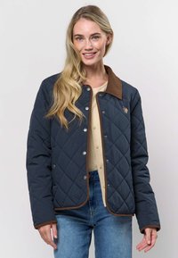 Šūta tumši zila jaka ar brūna korduroja apkakli un sprādzēm, ar noslēpumainu logotipu un kontrastējošu brūnu apmali gar malām.