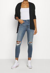 Czarny, ażurowy kardigan na biały top na ramiączkach, w połączeniu z postarzanymi niebieskimi skinny jeansami i białymi trampkami za kostkę.