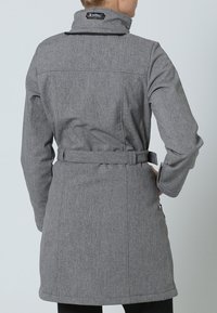 Manteau en tissu gris avec une taille ceinturée, doté d'un col et d'une finition texturée. Comprend un patch logo près du cou et des poches zippées.