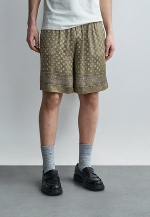 Vyras, apsirengęs alyvuogių žalius raštuotus šortus, pilkas kojines, juodus batelius ir baltą marškinėlį, stovintis ant pilkos grindų dangos prie vientisos sienos.