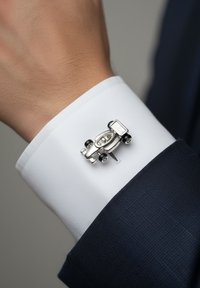Gemelos de color plateado con forma de coche de carreras, sujetos al puño de una camisa blanca, que contrastan con la tela de un traje azul oscuro.