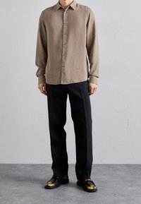 Camisa de manga larga marrón claro con botones, combinada con pantalones negros y zapatos negros brillantes con detalles amarillos. Textura suave y lisa.