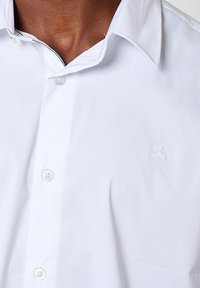 Witte gekraagde shirt met een gestructureerde afwerking, voorzien van een subtiel geborduurd logo op de borst en heldere plastic knopen.