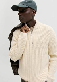 Pull en maille beige clair avec un col zippé, texture côtelée et manches longues, porté par-dessus un haut beige, avec une veste sombre drapée sur l'épaule.