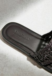 Sandale en cuir tressé marron foncé avec semelle plate et semelle intérieure cousue portant la mention « N. PREMIUM COLLECTION » sur un sol clair texturé.