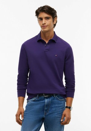 Junger Mann trägt ein lila langärmeliges Poloshirt und blaue Jeans, steht mit entspannter Haltung vor einem weißen Hintergrund.