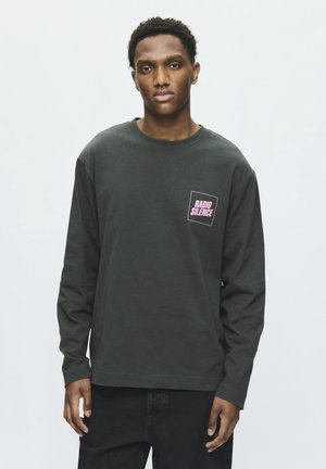 Jeune homme portant une chemise sombre à manches longues avec un texte rose "Radio Silence" sur la poitrine, debout devant un fond clair uni.