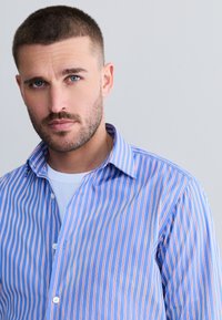 Plava i bijela prugasta košulja na kopčanje s crvenim naglascima, s ovratnikom širokog izreza, uskim krojem i glatkom teksturom.