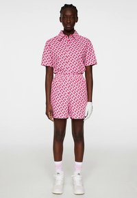 Rövid ujjú rózsaszín póló és rövidnadrág szett geometrikus fehér mintával. Galléros nyakkal és rugalmas derékkal rendelkezik, fehér tornacipővel párosítva.