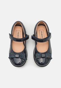 Námořnické lakované boty s kulatou špičkou, nastavitelným popruhem a třpytivou černou mašlí na přední straně. Hladká textura s lesklým povrchem.