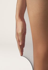 Gennemsigtige, nøgne strømpebukser hænger over en hånd, der hviler på et ben. Teksturen er glat med en gennemskinnelig finish, der fremhæver huden nedenunder.