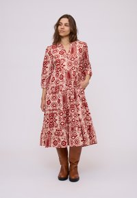 Floral patterned dress in red on a light background, with a V-neck, three-quarter sleeves, ruffled hem, and side pockets. Worn with brown knee-high boots.