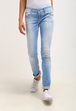Person iført lyseblå slim-fit jeans med opsmøgede bukseben og hvide sneakers, gående mod en ensfarvet baggrund.