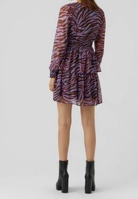 Mujer con un vestido de manga larga de estampado de cebra púrpura y marrón, con una falda de capas y botas negras hasta el tobillo, de espaldas.