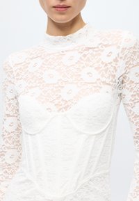 White lace bodysuit featuring a sheer floral pattern on long sleeves and a structured bodice with cups and vertical seams. High neck design.