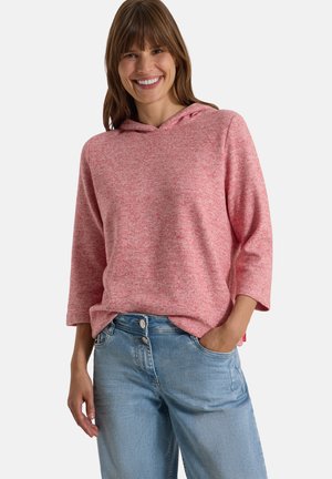 Femme souriante portant un haut à capuche rose à manches longues et un jean bleu clair, debout avec une main dans la poche sur un fond uni.